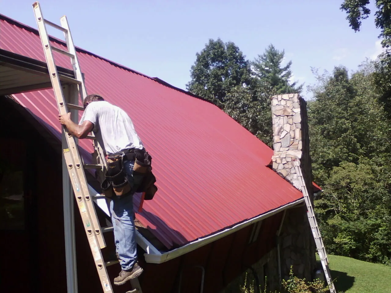 Roofer on a ladder beside a red metal roof and stone chimney — Soffit & Fascia Repair by Roofing Brothers in Cresson